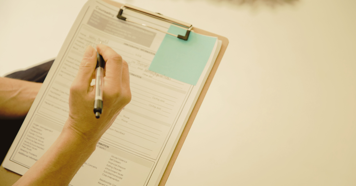 Close-up of a hand using a pen to complete a document with labeled sections on a clipboard, suggesting it could be a client form at an electrologist office.